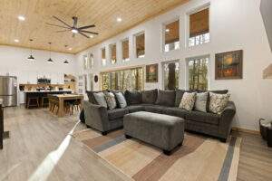 Spacious modern cabin interior featuring a gray sectional sofa, ottoman, and a large window offering a view of the Kentucky woods. The open-concept design showcases a kitchen and dining area under a warm wood ceiling, highlighting the craftsmanship of this custom built home by our cabin building company.