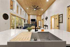 Bright, modern cabin interior featuring a white kitchen island with sink, open to a living area with a wood-paneled wall and large windows. A space to build your dream cabin with our construction company.