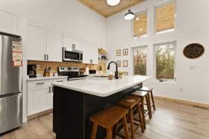 Modern cabin kitchen with white cabinets, a black island with wood stools, and large windows showcasing a wooded view. High ceilings and wood accents create a bright, inviting space for cabin building.