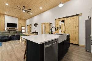 Bright, open-concept cabin interior with wood ceiling, white walls, and modern black kitchen island. Barn doors accent the space. A custom-built home design by our Kentucky cabin building company.