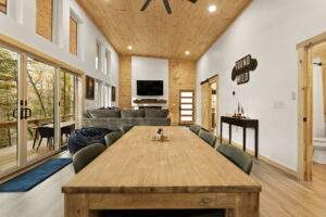 Spacious cabin interior with a long wooden dining table, leading to a living area with a gray sectional sofa and a wood-paneled wall with a TV. Large windows offer a view of the surrounding trees. "Found Wild" sign adds a rustic touch. Consider building with our cabin company!