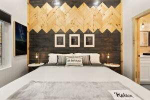 Cozy cabin bedroom featuring a custom wooden headboard with a chevron pattern above a dark wood panel, showcasing the craftsmanship of our cabin building company in the Red River Gorge. "Our Happy Place" pillow adorns the bed.