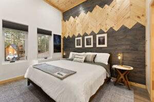 Cabin bedroom with a rustic wood accent wall in chevron pattern, white linens, and cozy decor. Sunlight streams through the windows, showcasing the serene cabin interior. Build your dream cabin with our custom construction services.
