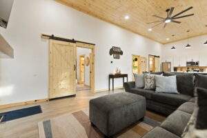 Cabin interior with light wood ceiling, white walls, and wood sliding barn doors. A gray sectional sofa and ottoman sit on a striped rug, with a "Found Wild" sign adding rustic charm. Cabin construction in Kentucky.