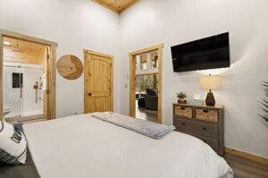 Cabin bedroom with rustic wood trim, a cozy bed, and a mounted TV. A doorway leads to a modern bathroom, showcasing cabin construction details in a Red River Gorge build.