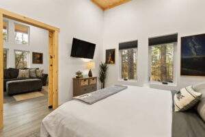 Bright, modern cabin bedroom with white walls, wood accents, and a view of the forest. A gray couch is visible in the next room. Build your dream cabin with our construction company!