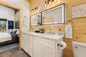 Bright, modern cabin bathroom featuring a double vanity with white cabinets and speckled countertops, illuminated by twin LED mirrors and rustic sconces against wood paneled walls. A glimpse into the bedroom shows a cozy bed. Build your dream cabin with us!