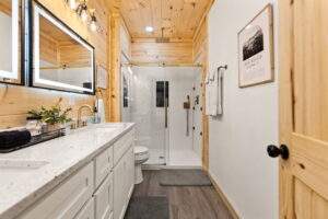 Bathroom with wood walls and white cabinets. A glass shower is adjacent to the toilet. A framed print reads "Red River Gorge." Build your dream cabin with our construction company.
