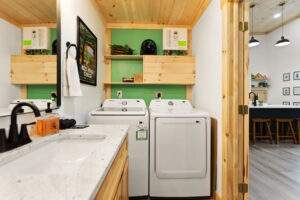 Bright, modern laundry room in a Red River Gorge cabin features a Samsung washer and dryer, light wood shelving against a green wall, and a view into a stylish kitchen with bar seating. The design highlights cabin construction.