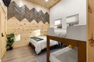A cozy cabin bedroom features wood paneling with a chevron accent wall, a neatly made single bed, and a bunk bed. Natural light streams through the windows, highlighting the room's warm, inviting atmosphere, perfect for a Red River Gorge cabin retreat.