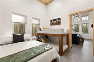 Cabin interior with a queen bed, bunk beds, and kitchenette. Light wood accents, white walls, and mountain art create a cozy vibe. Build your dream cabin with our construction company in the Red River Gorge.