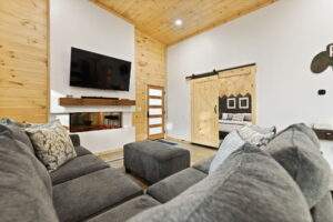 Cozy cabin living room featuring a gray sectional sofa, stone fireplace with a TV above, and a sliding barn door leading to the bedroom. Natural wood ceiling and accent wall add warmth. Build your dream cabin with us!
