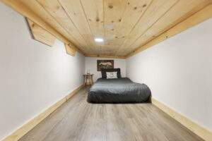 Cozy cabin nook featuring a low platform bed with gray bedding, nestled between white walls and a wooden plank ceiling. A small side table and decorative artwork complete the intimate space, showcasing a unique cabin design.