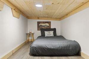 Cozy cabin bedroom featuring a low ceiling with wood planks, a single bed with gray bedding and a "Shine Bright!" pillow, and a small side table with a vase. Custom built home construction in Kentucky.