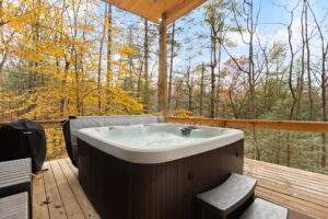 Hot tub on a wooden deck surrounded by fall foliage and trees. A covered grill sits nearby, promising relaxation and outdoor enjoyment in a custom built home.