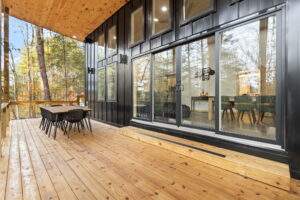 Modern cabin with a black exterior, large sliding glass doors, and a wooden deck. A dining table with black chairs is set on the deck, overlooking trees. Build your cabin with us in the Red River Gorge!