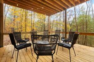 A cozy, covered porch with a wooden deck and modern black chairs arranged around a small table, overlooking a vibrant autumn forest. Perfect for relaxing in a custom built cabin by our Kentucky building company.