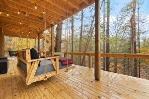 A cozy outdoor deck featuring a swinging daybed with a gray mattress and red plaid blanket. String lights hang from the wooden ceiling, creating a warm ambiance. The deck is surrounded by tall trees, showcasing the beauty of the Red River Gorge.