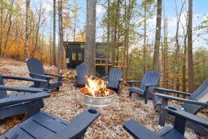 A cozy Kentucky cabin fire pit with Adirondack chairs. The fire blazes in a metal pit, surrounded by gravel and fall leaves. A modern black cabin stands in the background, ready for building by our construction company in Red River Gorge.