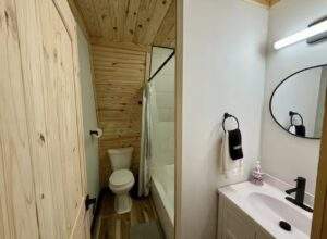 Cozy cabin bathroom featuring wood-paneled walls and ceiling, a toilet, a shower with a curtain, a white vanity with a black faucet, and a round black-framed mirror. Build your dream cabin with our construction company!