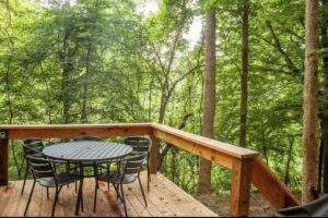 Wooden deck with a round table and six chairs overlooking a lush green forest. Perfect for a custom-built cabin in the Red River Gorge, constructed by our cabin building company in central Kentucky.