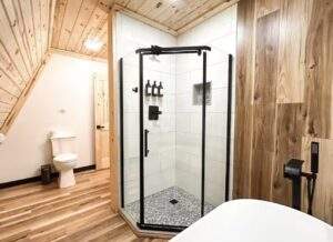 Rustic cabin bathroom featuring a modern glass shower with black trim, white subway tile, and a mosaic tile floor. Wood plank walls and ceiling add warmth. Toilet and freestanding tub visible.