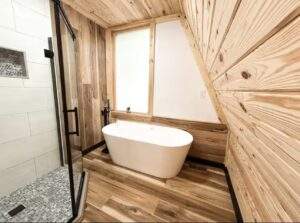A-frame cabin bathroom featuring a freestanding white tub beneath a window, next to a glass-enclosed shower with white subway tile and stone flooring. Wood paneled walls and flooring create a warm, natural aesthetic, showcasing a Red River Gorge cabin design.
