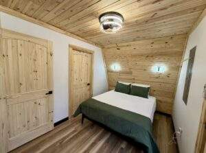 Cabin bedroom featuring wood paneled walls and ceiling, a bed with white sheets and green accents, and modern lighting. A cozy and rustic design by our cabin building company.