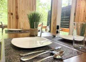 A modern dining table set for two in a cabin, featuring woven placemats, square white plates, silverware, and clear glasses. A small potted plant adds a touch of nature. Build your cabin with us!