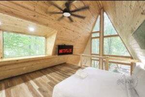 Cozy A-frame cabin interior with wood paneling, large windows showcasing a forest view, a bed, and a TV displaying Netflix. Consider building your custom cabin with our construction company in Red River Gorge, Kentucky.