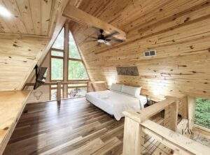 A-frame cabin interior featuring a queen bed, wood paneled walls, and a large triangular window showcasing the surrounding Red River Gorge scenery. Cabin built by our construction company in central Kentucky.
