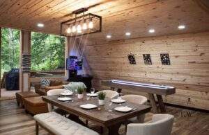 A beautifully constructed cabin interior features a dining area with a wooden table set for dinner, a shuffleboard table, and a cozy seating area, showcasing the craftsmanship of a custom-built home. Large windows reveal a lush, green forest outside.