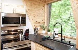 Modern cabin kitchen featuring stainless steel appliances, black countertops, and a large window overlooking lush greenery. A cozy and stylish space built by our cabin building company in Kentucky.