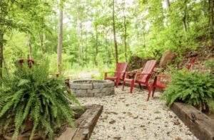 An outdoor fire pit area encircled by red Adirondack chairs, ferns, and gravel nestled in a lush Kentucky forest. Perfect for relaxing near a custom-built cabin or home in the Red River Gorge area.