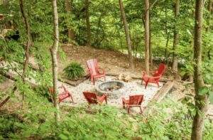 A cozy fire pit area in the woods, featuring red Adirondack chairs arranged around a stone fire pit. This inviting outdoor space, perfect for a custom-built cabin in Kentucky, is surrounded by lush greenery and trees.