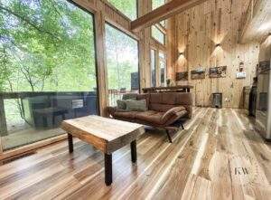 Cabin interior with wood paneling, large windows showcasing the forest. A brown couch and rustic wood coffee table sit on a wooden floor. Build your dream cabin with our Kentucky cabin building company.