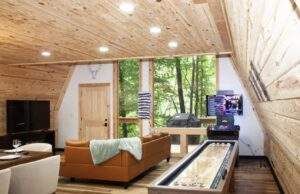 Cabin interior with wood paneling, featuring a shuffleboard table, leather couch, TV, and large windows showcasing a lush forest. Ideal space for construction of a custom cabin with our cabin building company in the Red River Gorge.