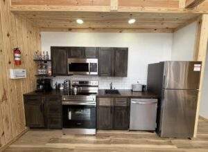 Cozy cabin kitchen featuring dark wood cabinets, stainless steel appliances, and light wood paneling. A fire extinguisher and first aid kit are mounted on the wall, emphasizing safety in this custom-built Kentucky cabin.