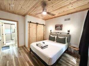 Rustic bedroom with wood-paneled ceiling, sliding barn doors, and a king-size bed. A modern bathroom is visible through an open doorway. Build your dream cabin in the Red River Gorge with our custom construction services.