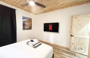Cozy cabin bedroom featuring a white bed with folded towels, a wall-mounted TV displaying Netflix, and a rustic wooden door. The room has a wood plank ceiling and flooring, creating a warm and inviting atmosphere. Consider our cabin building company for your custom home in Kentucky.