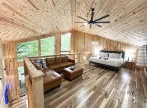Cozy cabin interior with wood walls, floors, and ceiling. A leather couch and ottoman sit near large windows overlooking trees. A bed with green headboard is on the right. Build your custom cabin with us in Kentucky!