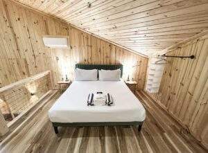 Cozy, light-filled cabin bedroom featuring a queen bed with crisp white linens and a green headboard. Wood paneled walls and ceiling showcase the craftsmanship of this Red River Gorge cabin, built by our custom cabin building company.
