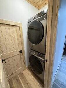 Stacked Samsung washer and dryer unit in a newly constructed laundry room. Natural wood trim complements the light wood flooring and door, highlighting the quality craftsmanship of this custom built home.