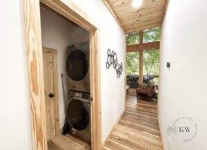 Hallway in a custom built cabin featuring a stacked washer and dryer in a closet, light wood floors, and a window wall showcasing the Red River Gorge scenery. Build your dream cabin with our construction company.