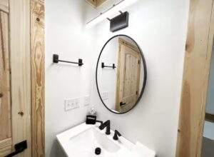A modern bathroom with a white sink, black faucet, and round mirror. The design features natural wood accents, highlighting construction and cabin building.