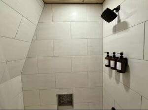 Modern shower with large rectangular white tiles, a dark rainfall shower head, and mounted brown bottles. Cabin-style design with wood ceiling. Consider our cabin building company for your construction needs.