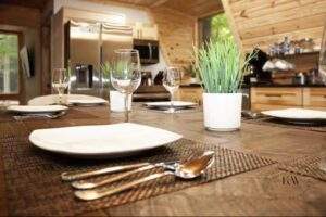 Cozy cabin dining room featuring a rustic wooden table set with white plates, silverware, and wine glasses. Green plants add a touch of nature. Perfect for a custom-built home by our cabin building company in the Red River Gorge.