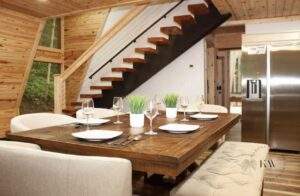 A modern dining room with a rustic wood table set for six, featuring a floating staircase and light wood paneling, showcasing a cabin-style interior design ideal for building your dream home with our construction company.