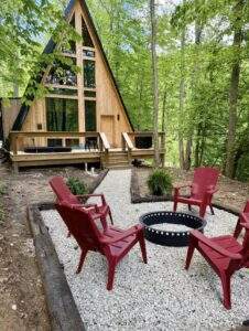 A-frame cabin nestled in the woods with a stone fire pit and red Adirondack chairs. A perfect scene to inspire building your dream cabin with our company.