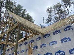 Cabin construction in progress: the wooden frame of a Red River Gorge cabin rises against a cloudy sky, showcasing the skeletal structure and Lowe's house wrap. Builders work on the roof rafters, highlighting the custom built home process.